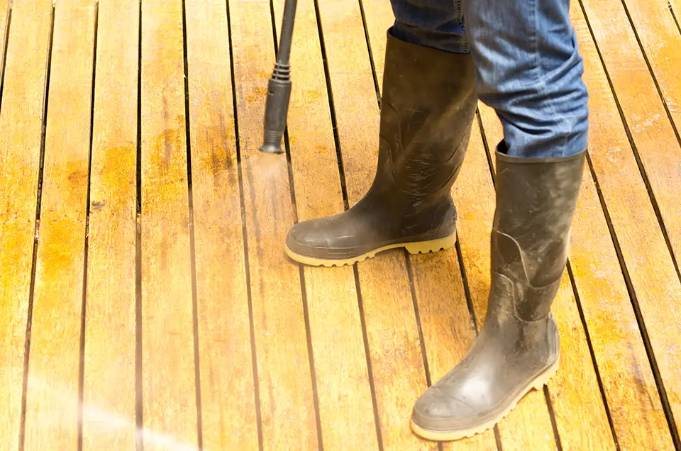Man wearing rubber boots using low water pressure cleaner on wooden terrace surface.